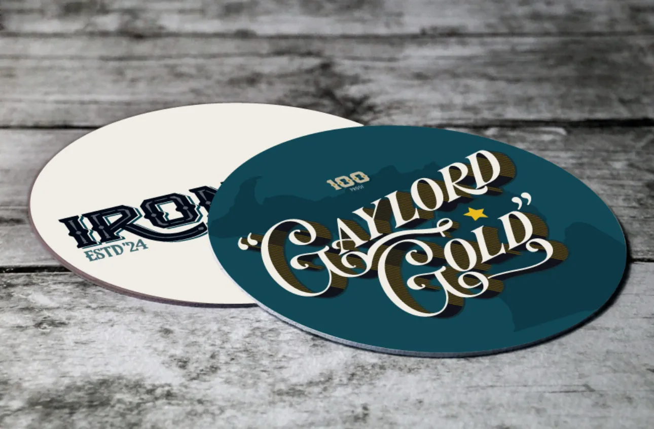 Iron Pig Bourbon Gaylord Gold Round Coasters, one cream Iron Pig ESTD '24 and one teal Gaylord Gold, on weathered wood